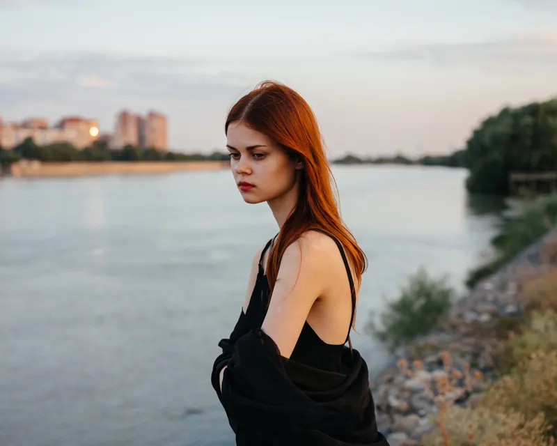 Рыжеволосая женщина в черном платье стоит у реки на природе