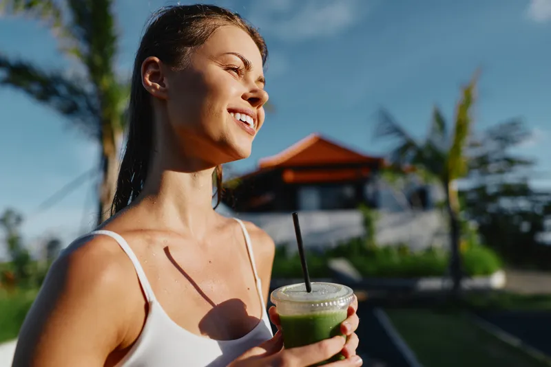Smiling woman in a white top enjoying a green smoothie outdoors on a sunny day, with lush background and palm trees, capturing a lifestyle moment of wellness and self care.