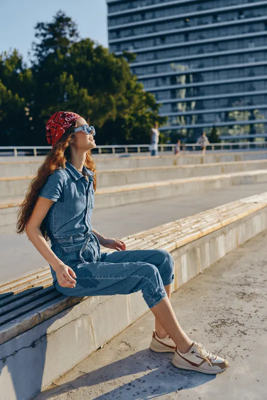 Lifestyle woman in modern boho-western denim street style relaxes outdoors wearing a red bandana and sunglasses, captured with warm film color tones in an urban setting.