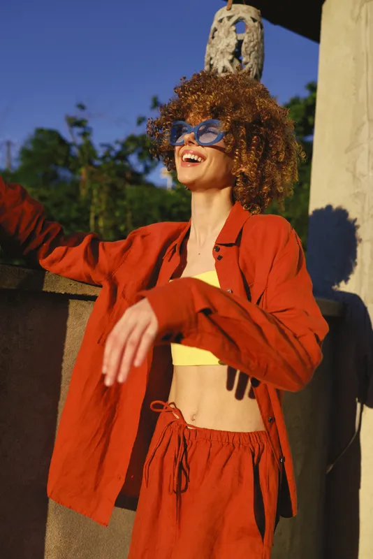 Bright woman with curly hair wearing a red orange jacket, cropped top, and sunglasses, posing with arms raised against a sunny outdoor backdrop, conveying energetic summer confidence