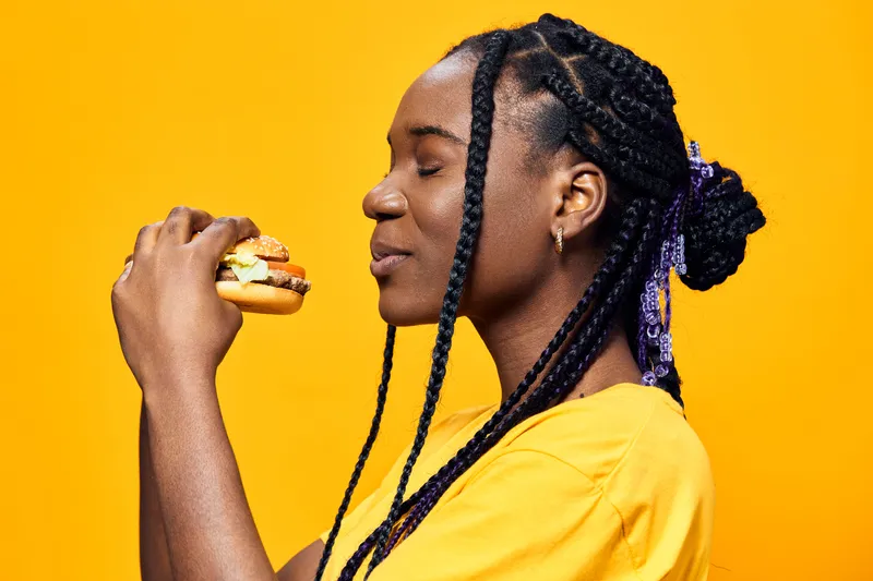 Young woman enjoying a colorful burger against a bright yellow background, highlighting healthy eating and vibrant emotions