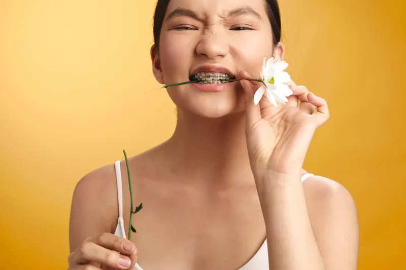 asian woman smiles softly while biting a flower stem, playful portrait in a bright studio against a warm backdrop, highlighting youthful charm and lighthearted style.