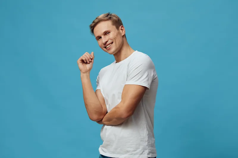 Smiling young man with blonde hair wearing a white t-shirt poses confidently on a plain blue background studio portrait. People lifestyle concept