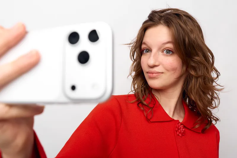 Young woman with curly hair wearing red sweater takes selfie with white smartphone on white background. Concept of social media, technology, and casual lifestyle portrait.