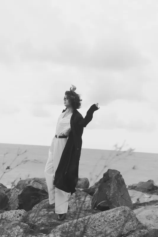 Woman stands on rugged rocks along the windy coast, arms raised, long coat flowing, sea breeze, black and white scene, sense of release, exploration and solitude by the water.
