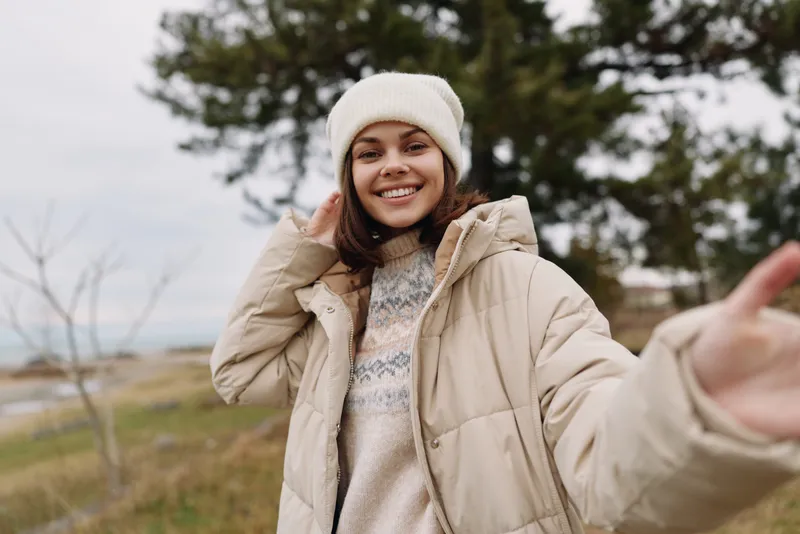 Woman outdoors in a warm coat and beanie, smiling in a windy countryside landscape. Casual fashion, cozy vibe, and relaxed portrait captured with natural light in an open field.