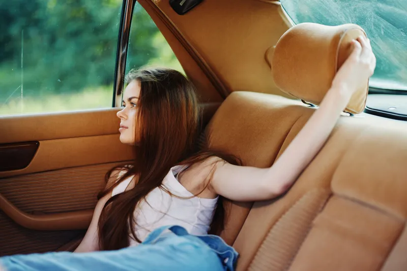 Young woman with long hair in a casual outfit, resting in a vintage car, expressing a sense of relaxation amidst a natural background, perfect for lifestyle and travel themes.