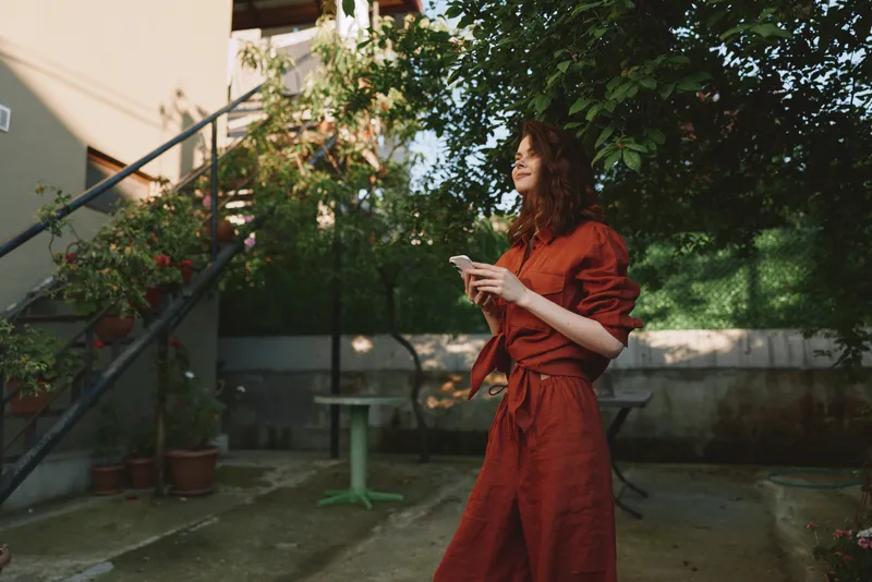 A woman in a red dress stands outdoors, holding a smartphone, smiling softly. Sunlit garden and potted plants frame a relaxed lifestyle moment perfect for everyday connectivity