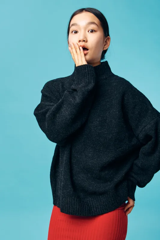 Young woman in black sweater and red skirt with surprised expression covering mouth against blue background. Studio portrait, casual clothing, emotion and outfit details.