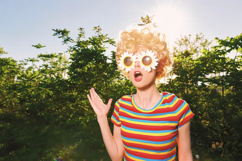Cheerful woman outdoors in a striped shirt with flower sunglasses, smiling with open mouth, sunlit moment, relaxed pose, retro film color mood, lush greenery background