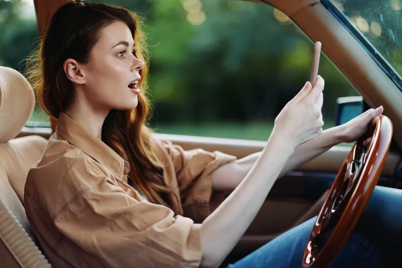Young woman driving a car and using a smartphone, expressing surprise and excitement in a casual outfit, surrounded by lush greenery.