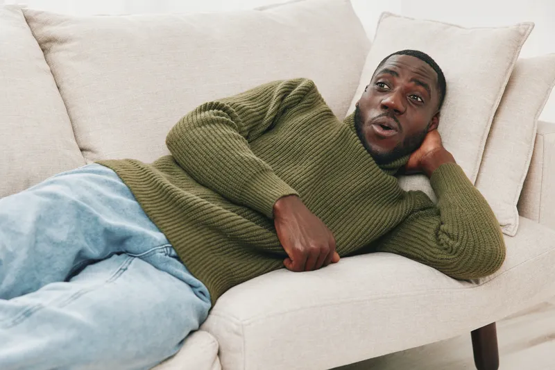 Young African man relaxing on a beige sofa, wearing a green sweater and jeans, expressing surprise and joy in a cozy indoor setting