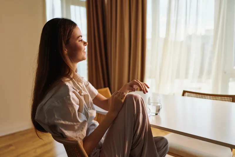 Woman home morning sitting by window smiling and relaxed at dining table with glass of water, casual outfit and soft natural light, cozy interior and peaceful domestic moment.
