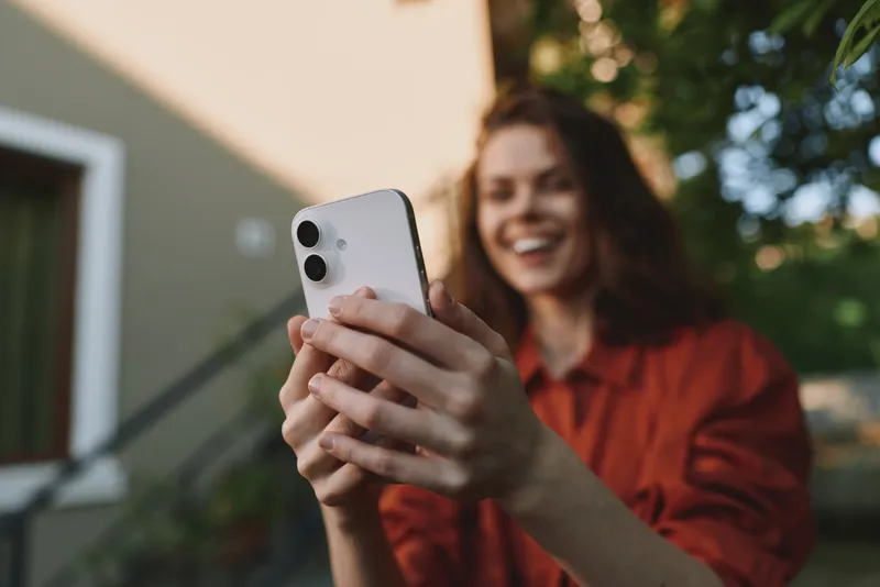 A smiling woman outdoors captures a selfie with a smartphone, wearing a casual red top. Bright natural light highlights a relaxed, carefree lifestyle moment