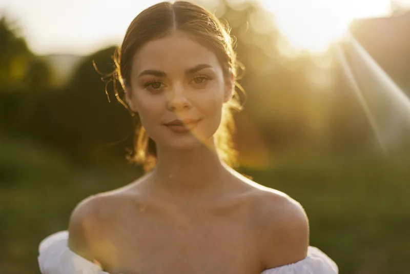 Beautiful young woman in a white off shoulder dress, smiling softly against a warm sunset background, evoking feelings of serenity and elegance