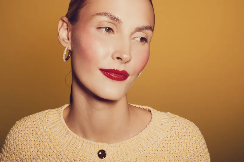 Close-up portrait of a woman with red lipstick and hoop earrings wearing a knitted sweater. Soft lighting highlights her smooth skin and serene expression on a warm beige background.