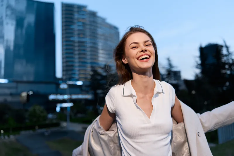 Confident woman in business attire smiling outdoors with modern city buildings in the background, exuding positivity and professionalism.