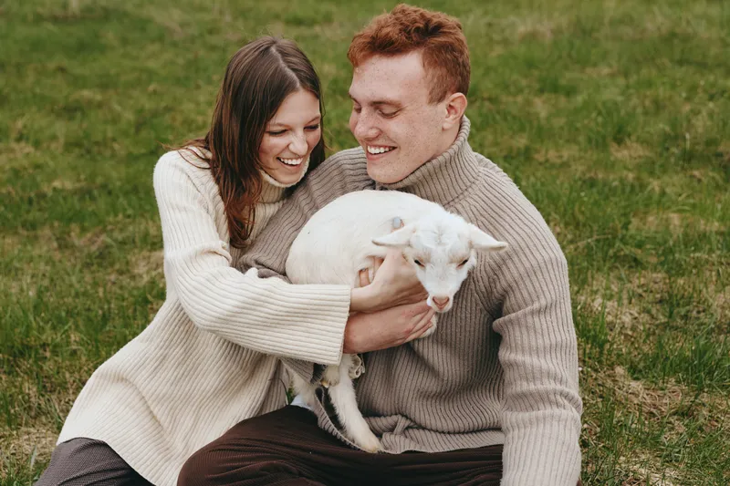 Smiling couple in knit sweaters cuddle a white goat kid outdoors on green grass, capturing a warm lifestyle moment of companionship, joy, and rural simplicity