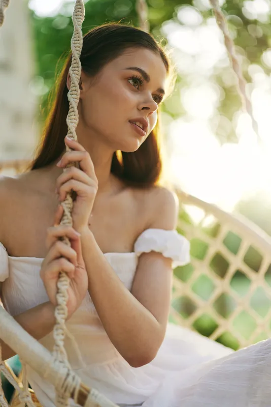 Young woman in a white dress sitting on a swing, enjoying a sunny day, reflecting tranquility and beauty in a natural outdoor setting