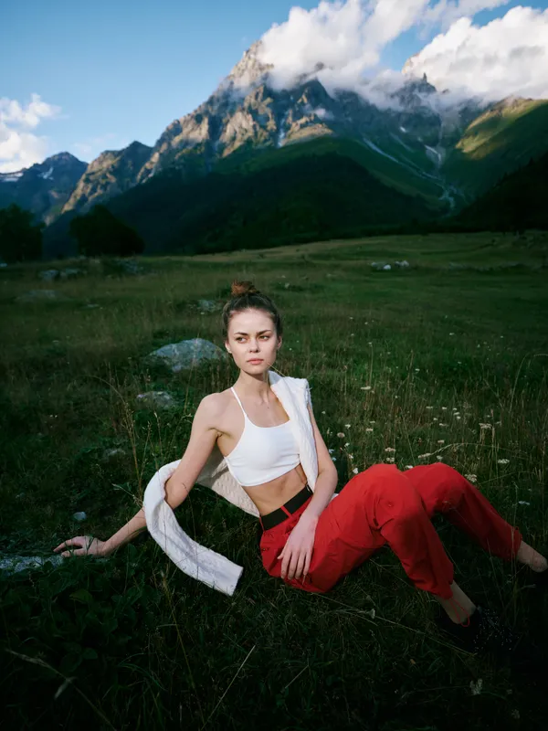Woman lounging in a meadow with mountains in the distance, casual white top and red pants, relaxed pose under open sky among wildflowers