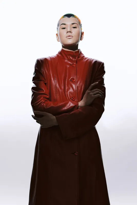woman in red leather coat with folded arms posing confidently against white background in studio lighting, stylish fashion portrait of strong young woman