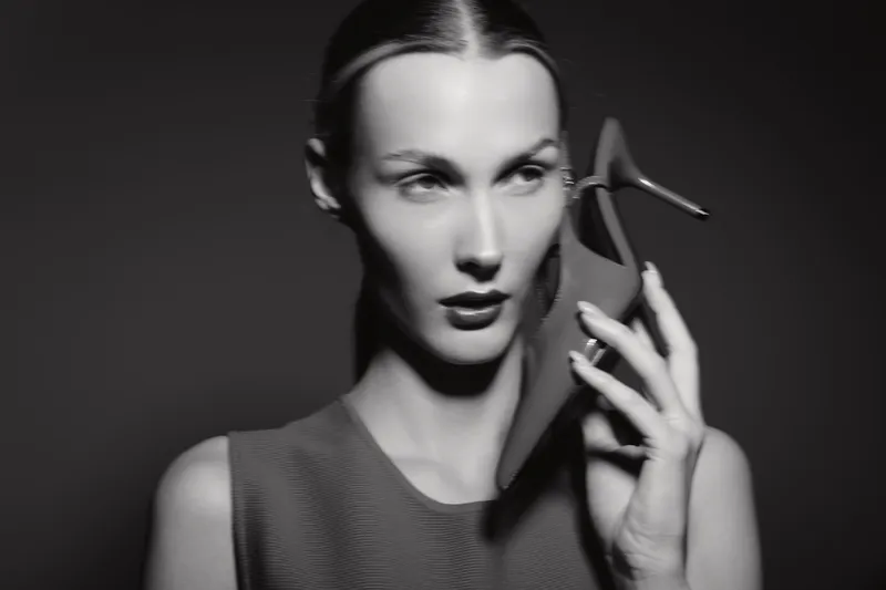 White woman with pale skin holds high heel shoe near face in moody black and white studio portrait. Concept for fashion, elegance, and style.