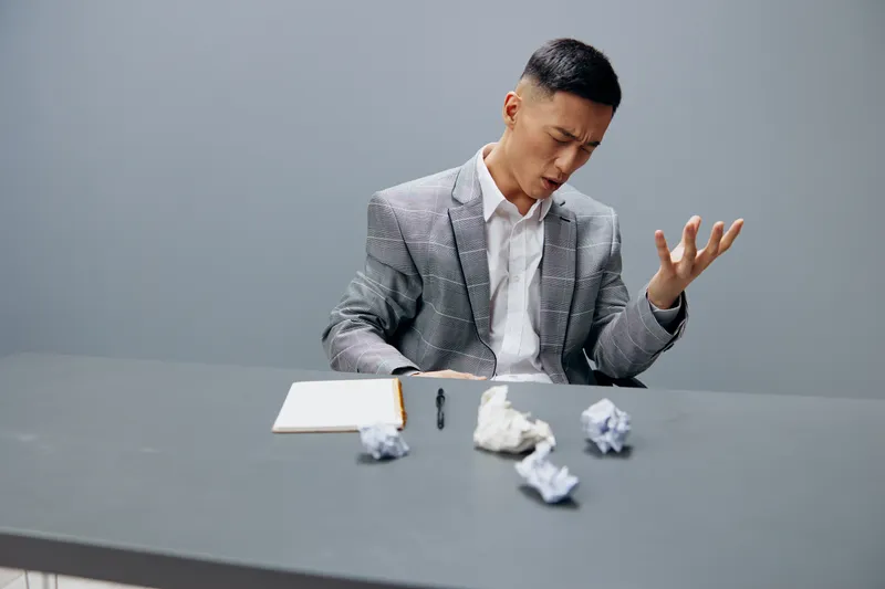Работник в костюме сидит за столом, на его лице эмоции, рядом смятая бумага, изолированный фон