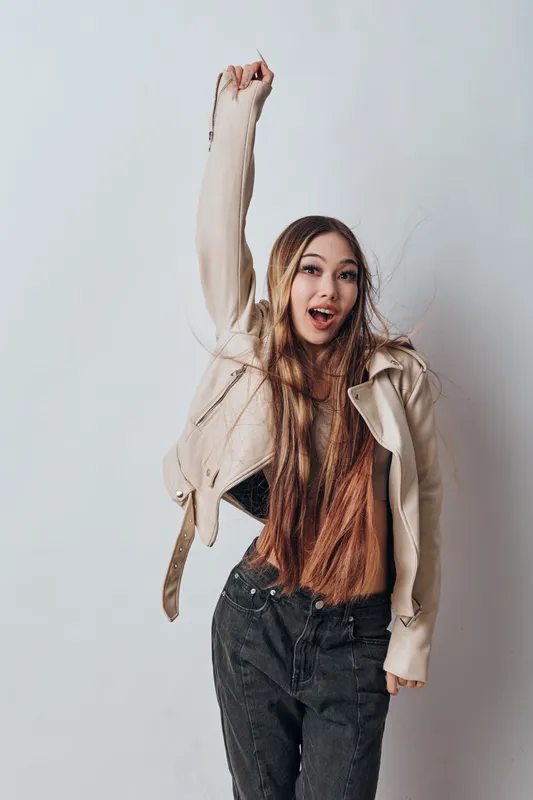 Young woman with long hair raising her arm up wearing a beige jacket and black pants posing against light background with energetic facial expression and casual style.