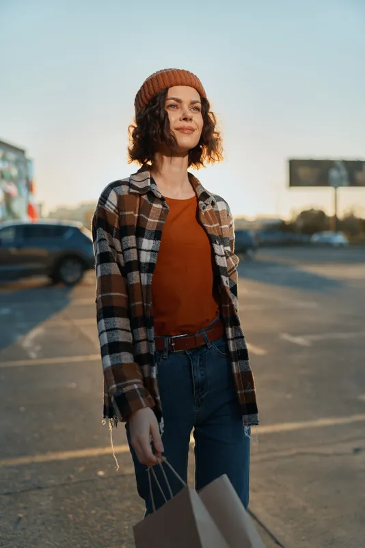Woman shopping with paper bags and smile in urban parking under warm sunlight, wearing beanie and plaid shirt; authenticity, candid lifestyle, golden hour glow and emotional storytelling mood.
