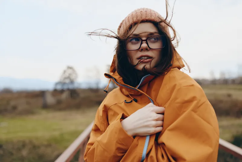 Woman lifestyle outdoors raincoat hat glasses wind countryside portrait autumn. Female traveler in orange raincoat adjusts zipper, beanie and glasses, windy rural field background, natural daylight.