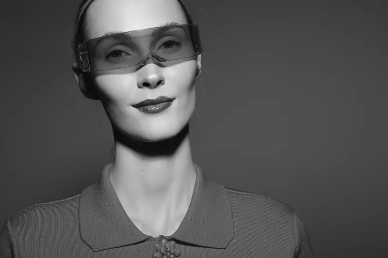 Caucasian woman with fair skin wears transparent sunglasses and textured top, posing in studio with calm confident expression.