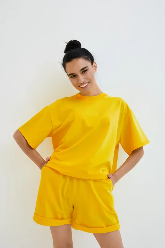 smiling woman in yellow casual outfit stands against white background with hands on hips, showing happiness and relaxed summer style in bright warm colors
