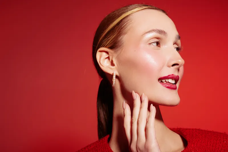 Young woman with shiny skin and red lips looks sideways on red background. She touches her face gently, wearing red sweater and gold earrings. Concept of beauty, skincare, and makeup.