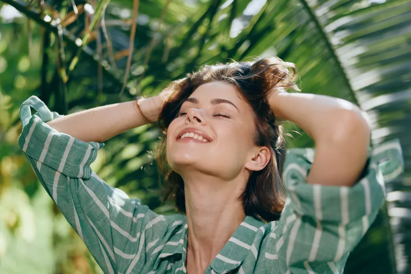 Smiling woman outdoors under bright sunlight, embracing a relaxed moment among lush tropical foliage and a striped shirt.