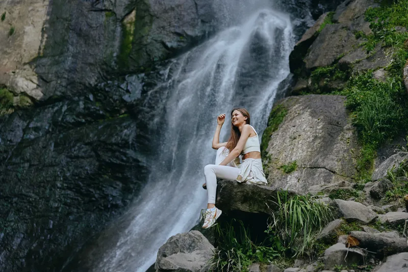 young woman sitting on rock near waterfall in nature, dressed in white activewear, enjoying peaceful outdoor adventure and fresh air during daytime.