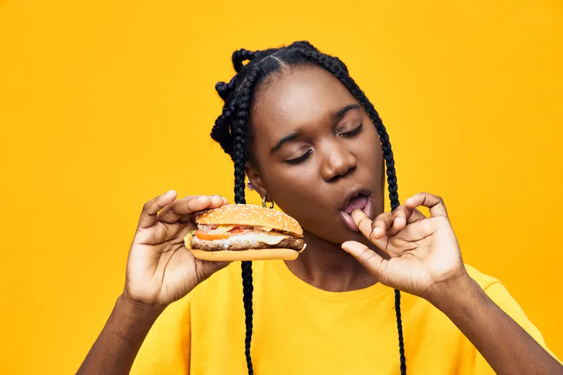 Happy Black woman enjoying a delicious burger against a vibrant yellow background, showcasing a fun and playful moment of indulgence in food
