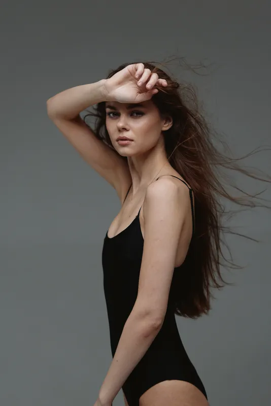 Young woman posing in black bodysuit with windblown hair on gray background, confident and elegant expression