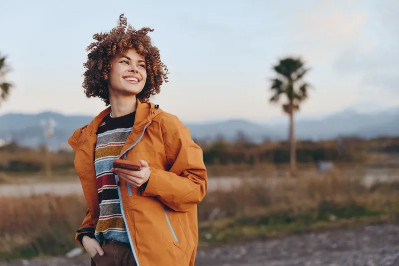 Woman lifestyle outdoors, smiling, curly hair, orange jacket, denim sweater, walking on rural path near field, meadow grass, mountains, palm trees, warm sunlight, casual leisure, adventure