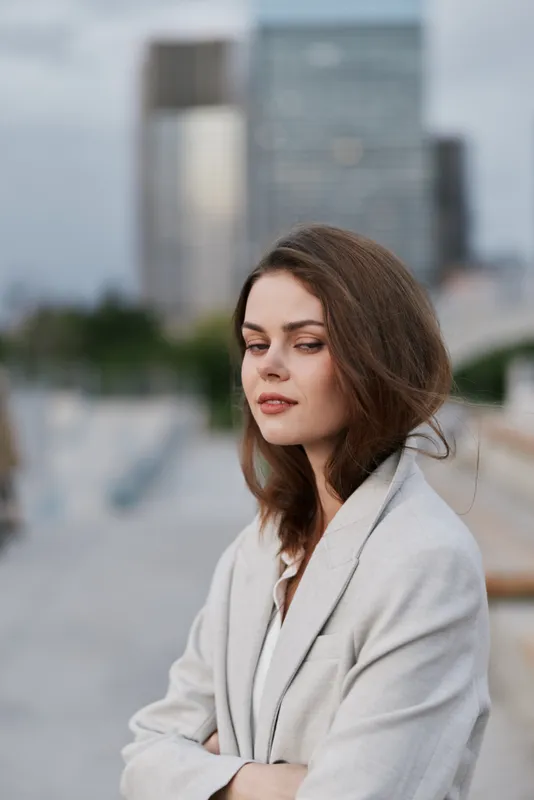 confident woman in light blazer with contemplative expression against urban cityscape background, soft natural colors, fashion concept