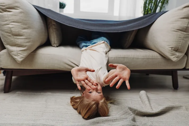 Playful child upside down under sofa blanket reaches hands forward with excited expression, cozy living room setting with soft carpet and natural light. Concept of childhood fun and imagination.