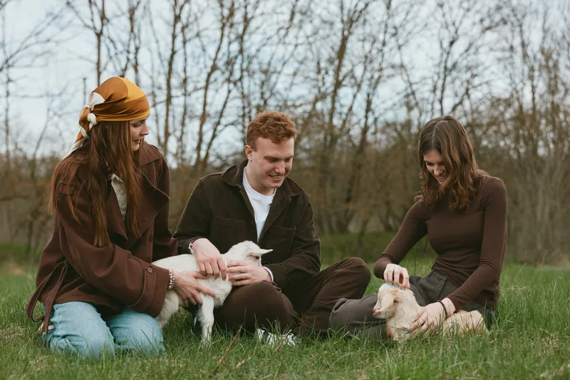 Three friends outdoors on a grassy field with a playful goat kid, sharing a gentle moment of companionship and care. Warm natural light highlights laughter, bonding, and rural lifestyle in a