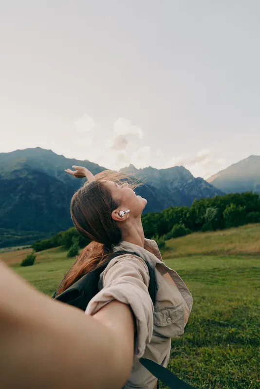Young woman with braided hair wearing earbuds holds hand and looks up while standing in green field with mountains in background under clear sky.