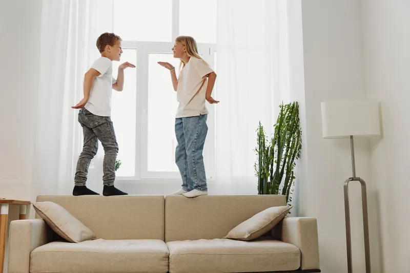 Two playful children standing on beige sofa in bright living room, making funny faces and gestures, enjoying carefree indoor fun together.