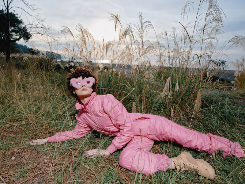 Woman in a pink jumpsuit lying in tall grass outdoors, wearing heart shaped pink sunglasses, playful and surreal portrait in a natural setting with soft light and open sky.