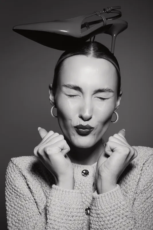Young white woman with pale skin and dark hair makes a playful face with closed eyes and puckered lips. She wears a knit sweater and balances a high heel shoe on her head in studio.