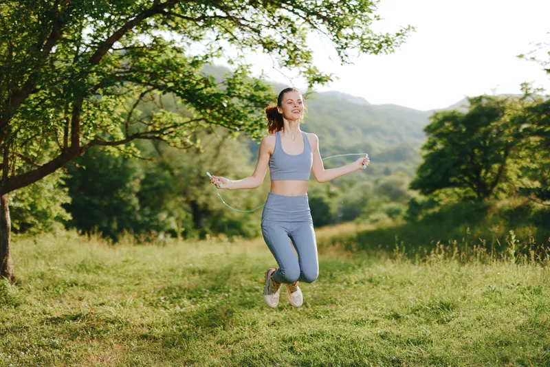 Young smiling woman jumping with a skipping rope in a green park, wearing activewear, embodying joy and health in a sunny outdoor setting