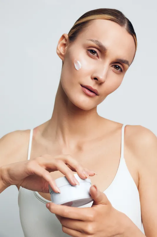 Young woman applies moisturizing cream on her face with gentle expression isolated on white background, skincare concept with natural beauty and healthy skin care routine.