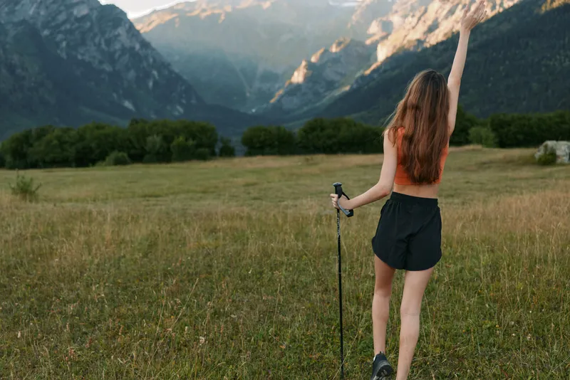 A young hiker stands in a grassy meadow with majestic mountains in the distance. She raises a hand in triumph, wearing a sleeveless top and shorts.