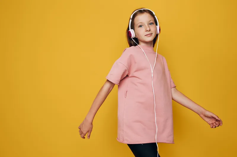 Girl wearing pink dress and headphones listening to music with relaxed pose on yellow background. Concept for leisure, youth, technology, and entertainment.
