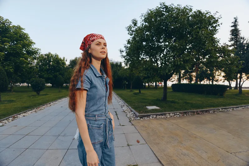 Lifestyle woman in modern boho-western denim street style wearing a red bandana, walking outdoors with natural greenery background, film color tone ambiance.